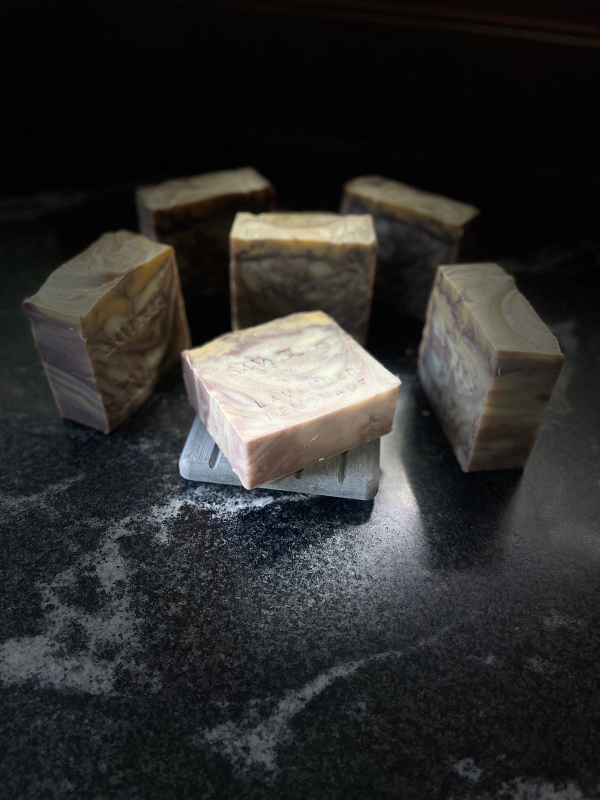 Five square bars of artisanal soap with marbled beige, brown, and cream swirls, arranged on a dark textured surface with one bar resting on a small gray slated soap dish