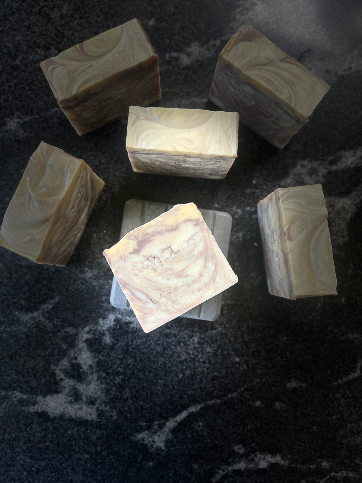 Six square bars of artisanal soap with marbled swirls of cream, beige, and soft purple, arranged on a dark marbled surface.