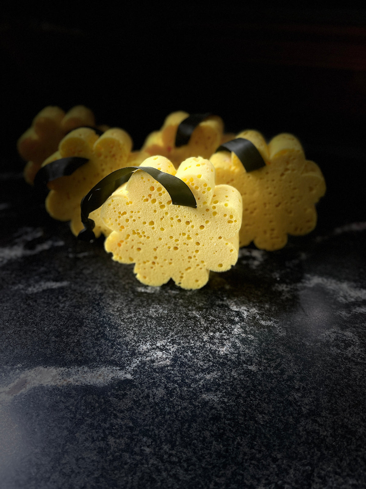 Several yellow soap sponge with black ribbon handles arranged on a dark textured surface