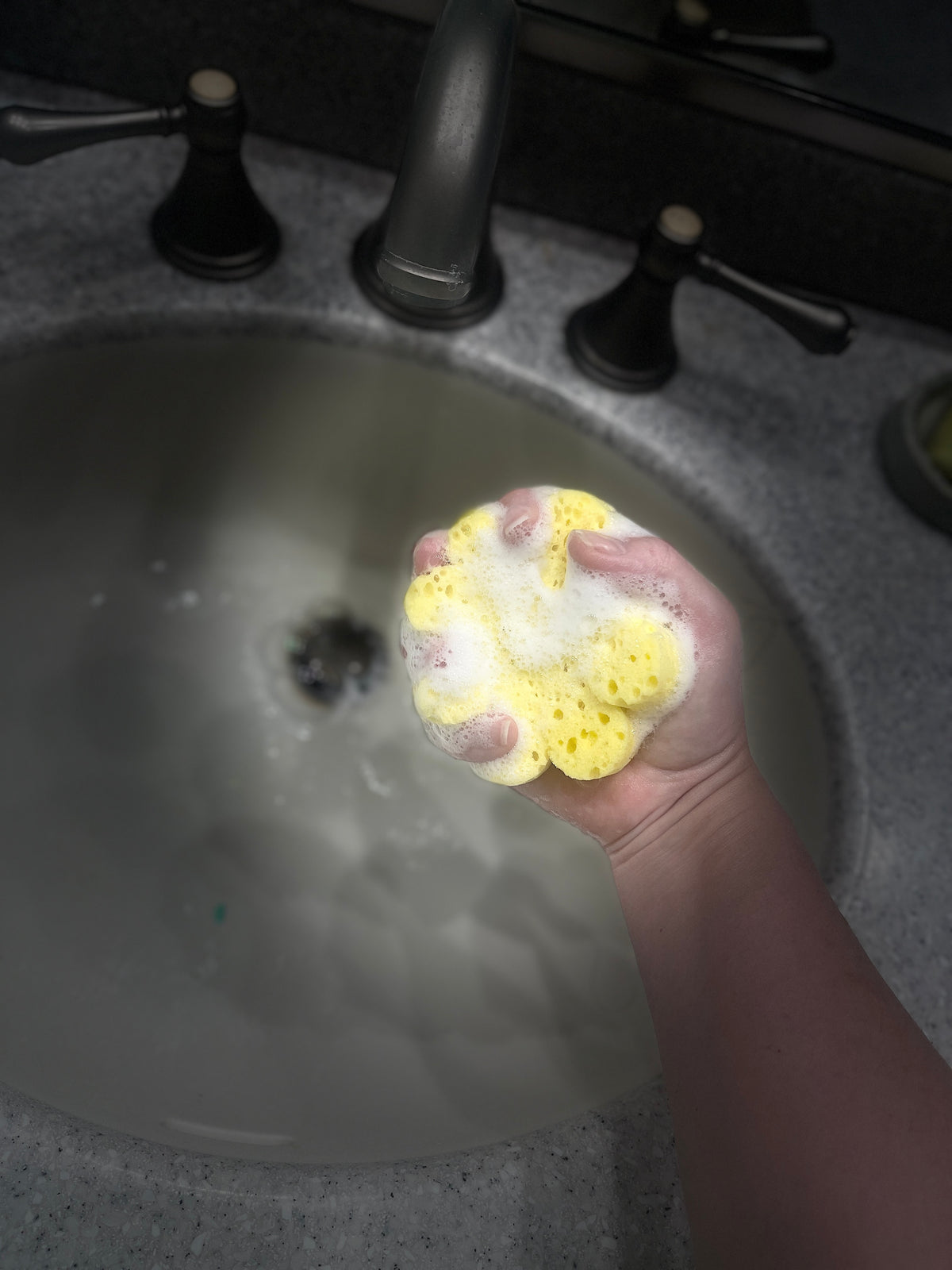 A hand holding a yellow sponge covered in white soap foam above a bathroom sink with dark faucet handles.