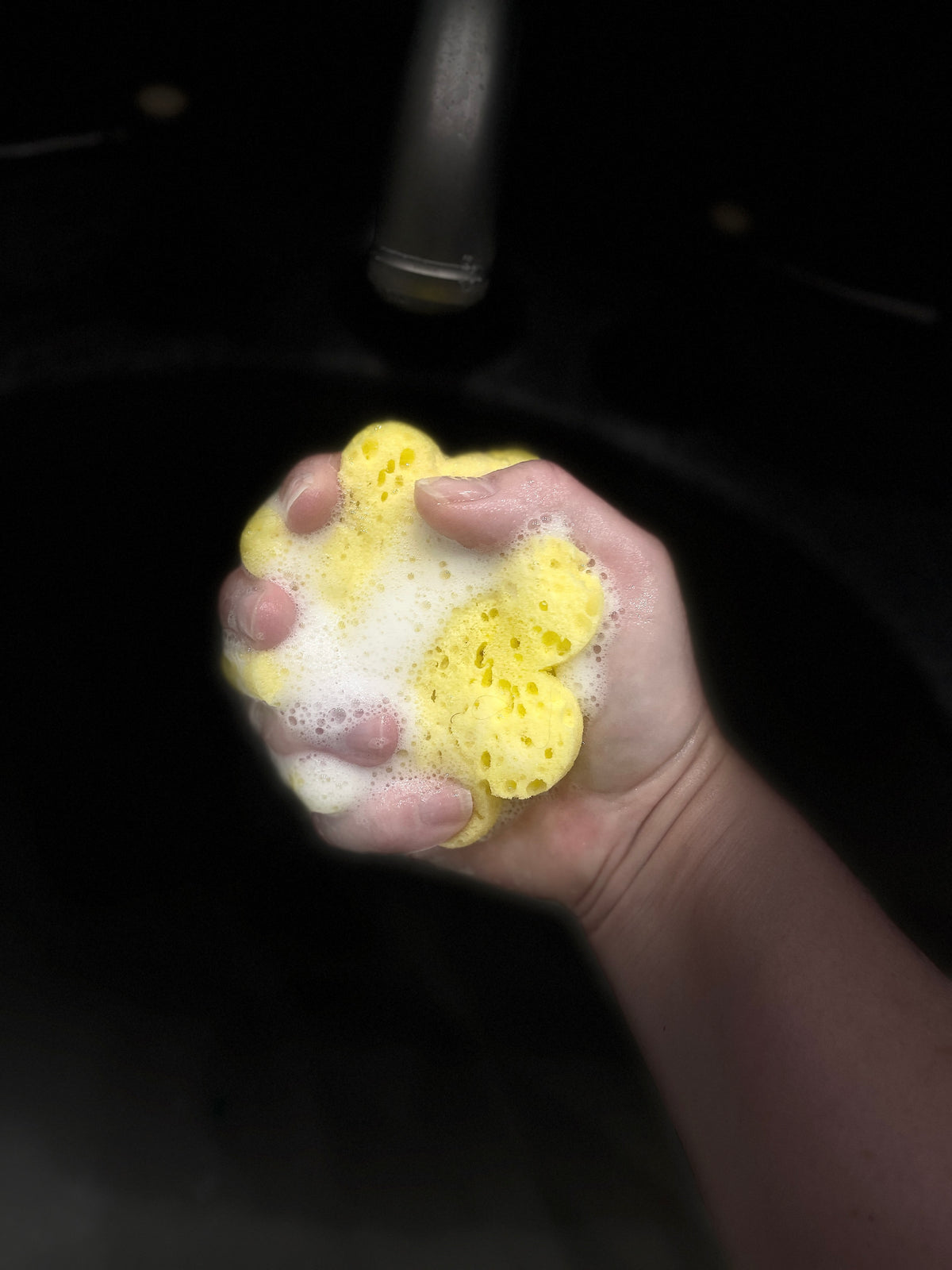 A hand squeezing a yellow sponge covered in white foamy soap over a dark sink with a faucet visible in the background.