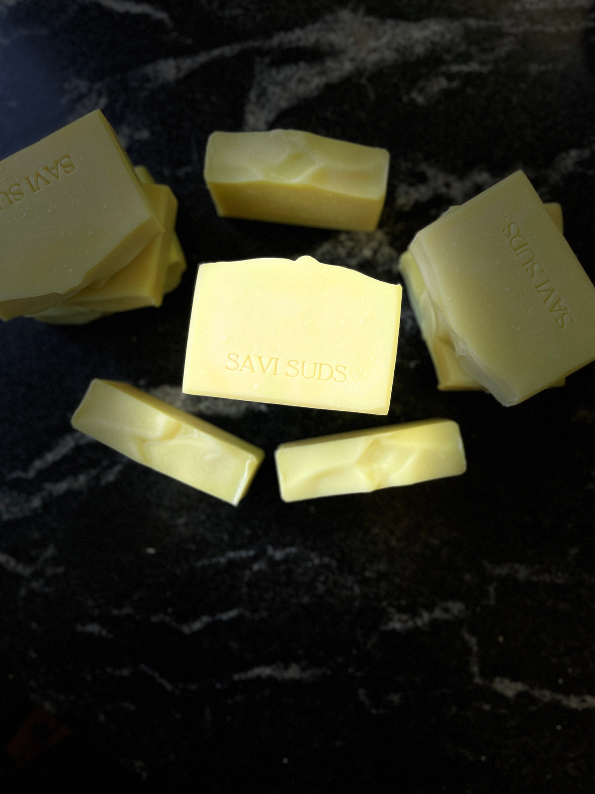 Several pale yellow bars of Savi Suds soap arranged on a dark marbled surface, with the brand name stamped on the tops.