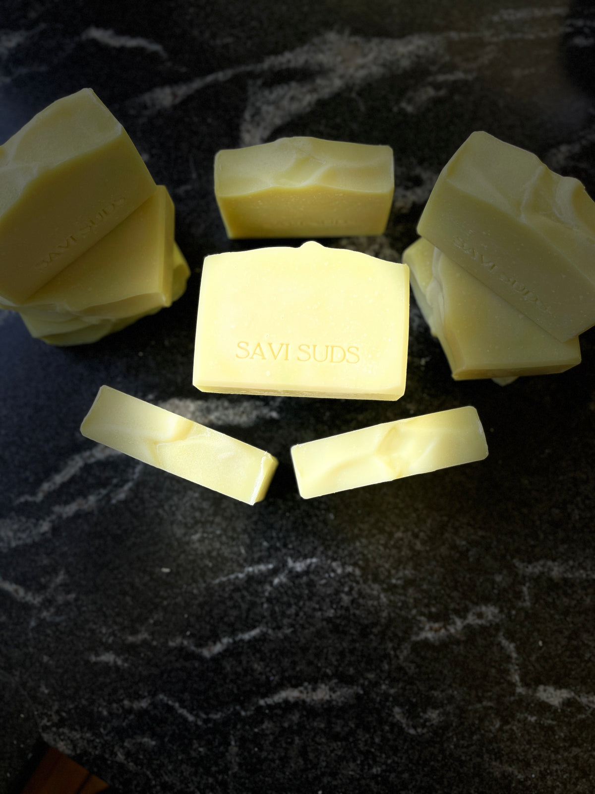 Several pale yellow bars of soap arranged on a dark marbled surface, with the center bar stamped with the brand name SAVI SUDS.