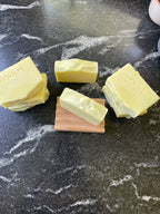 Several pale yellow handmade soap bars arranged on a dark marble surface, with three soaps stacked and one standing upright on a small wooden block. The brand name 'SAVI SUDS' is stamped on two soaps.