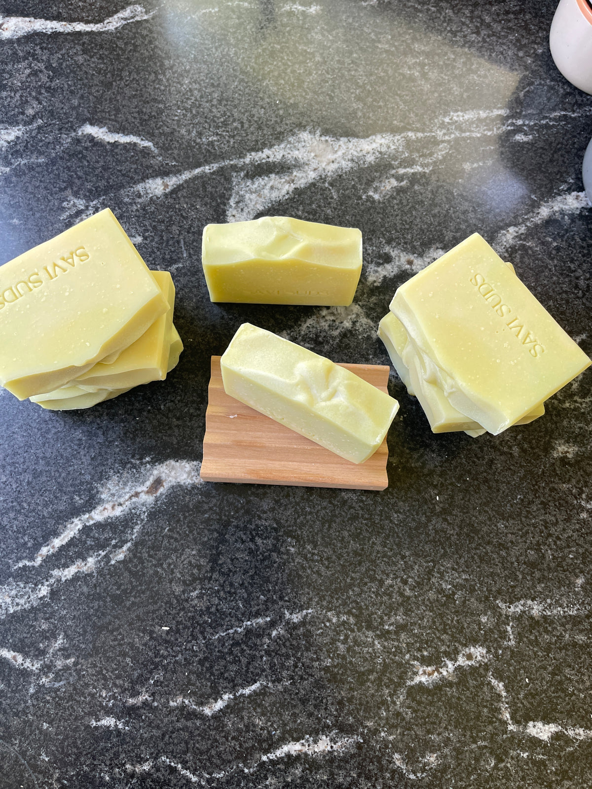 Several pale yellow handmade soap bars arranged on a dark marble surface, with three soaps stacked and one standing upright on a small wooden block. The brand name 'SAVI SUDS' is stamped on two soaps.