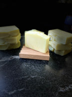 A pale yellow bar of soap stamped 'SAVI SUDS' displayed on a small wooden stand, with additional stacked soap bars in the background on a dark surface.