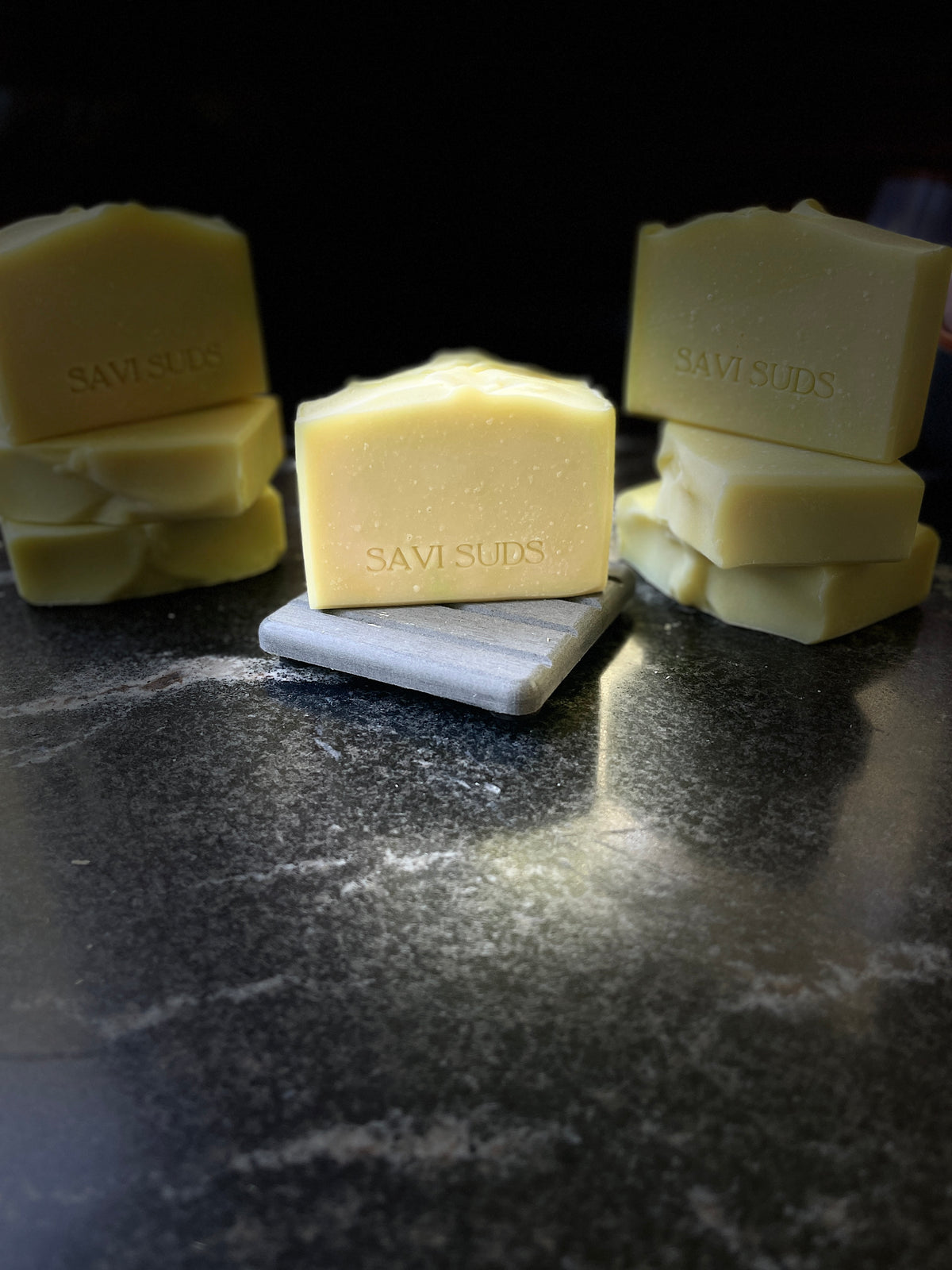 Several bars of Savi Suds handmade soap in pale yellow, arranged on a dark marble surface with a small gray slate tile under the center bar. The brand name 'SAVISUDS' is stamped on the faces of the bars.