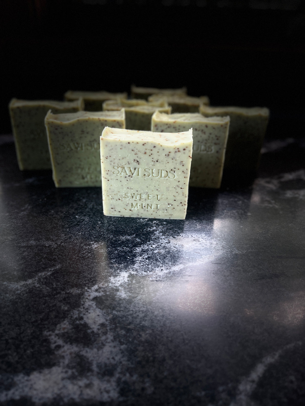 A handmade bar of soap labeled 'Savi Suds' stands in the foreground on a dark, reflective marble-like surface, with several matching bars blurred in the background.