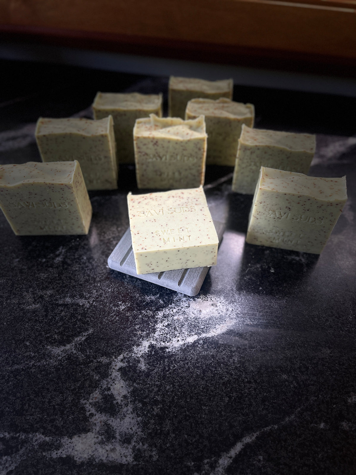 Several handmade square bars of soap in a pale green hue with small speckles, arranged on a dark marble-like surface with one bar resting on a small white tray.