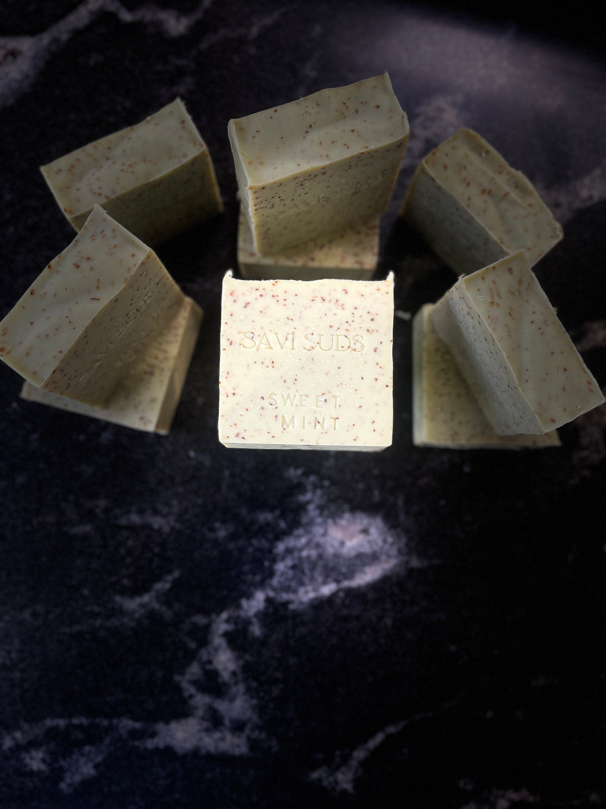  Multiple square bars of soap arranged on a dark marble-like surface, with one bar centered in front and embossed with the text 'SAVI SUDS'. The soaps are a pale green with small speckles from ground peppermint, and the background is a very dark black marble surface.