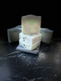 Three stacked handmade soap bars with visible botanical inclusions, featuring a front bar stamped with 'SAVISUDS'. The soaps have a creamy off-white color with subtle green and lavender swirls.