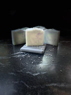 Three handmade square bars of soap in off-white, pale green and purple marbled soap, with one bar in the center resting on a small gray stone dish. The center bar is stamped with the brand name 'SAVISUDS'.