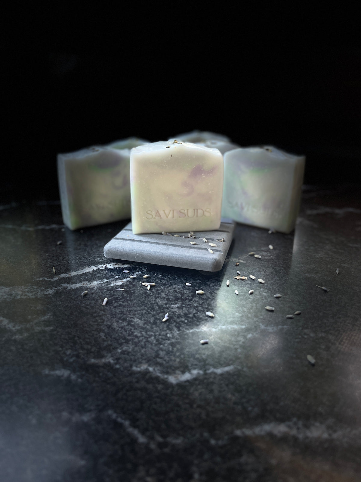 Three handmade square bars of soap in off-white, pale green and purple marbled soap, with one bar in the center resting on a small gray stone dish. The center bar is stamped with the brand name 'SAVISUDS'.