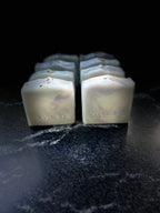 Eight square bars of Savi Suds soap arranged in two rows on a dark, textured surface against a black background.