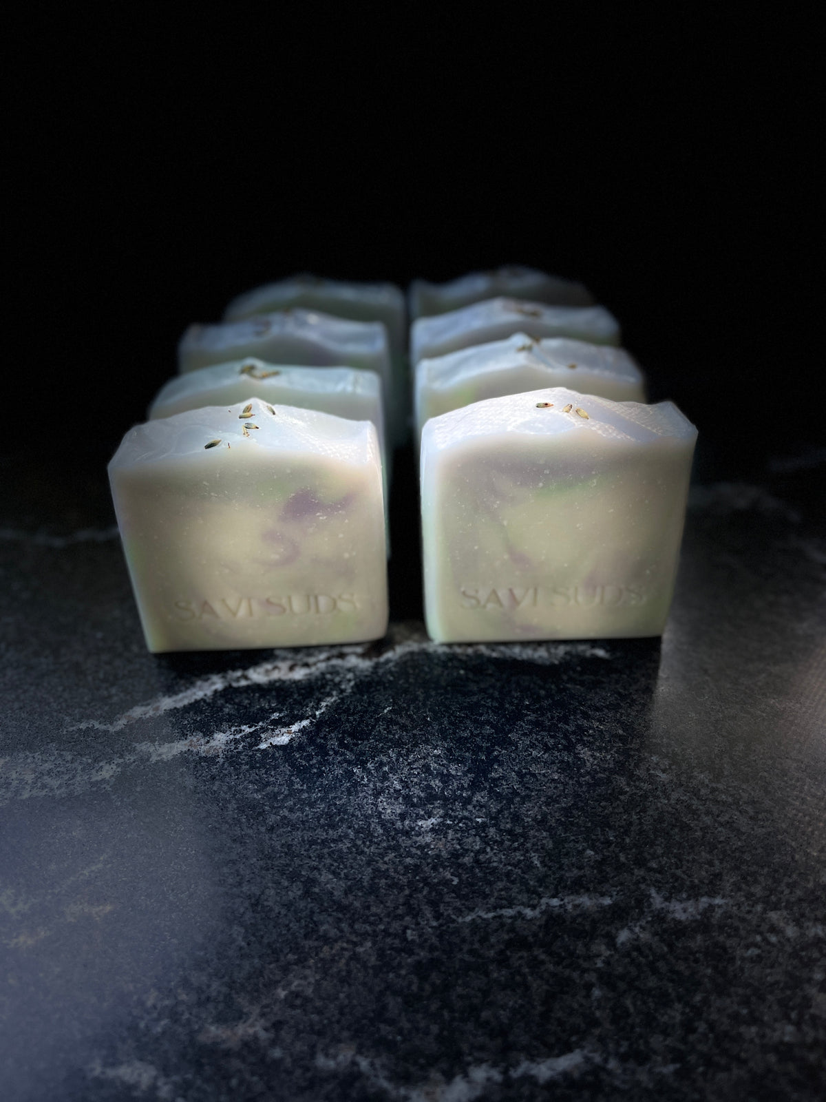 Eight square bars of Savi Suds soap arranged in two rows on a dark, textured surface against a black background.
