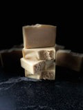 A stack of handmade soap bars in light beige and cream tones with oatmeal sprinkled on top of the bars. SAVI SUDS is stamped on the front of the top bar, set against a dark black background.