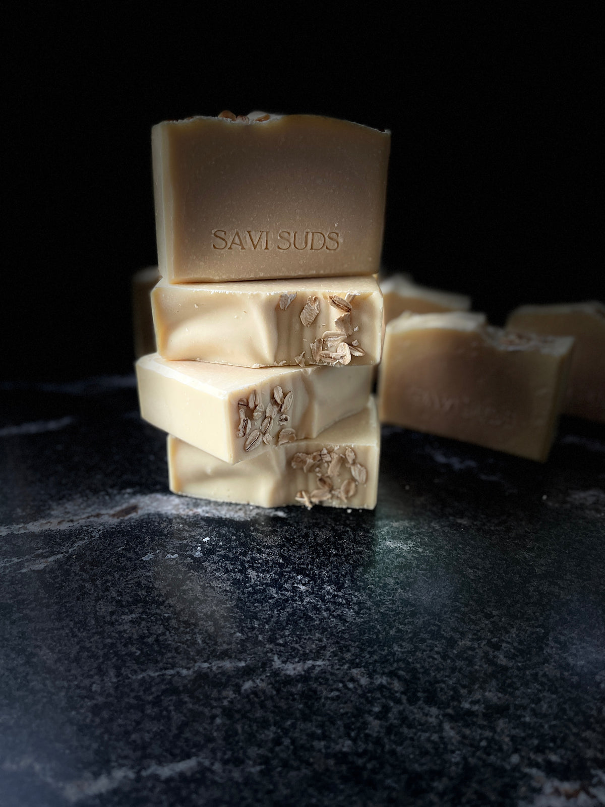 Four bars of Savi Suds soap stacked on a dark marble surface against a black background. The soap is a light cream color with oatmeal sprinkled on top