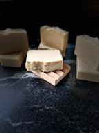 Several bars of handmade soap in warm cream and beige tones, with one bar laying on a wooden soap dish in front. They are arranged on a dark stone surface