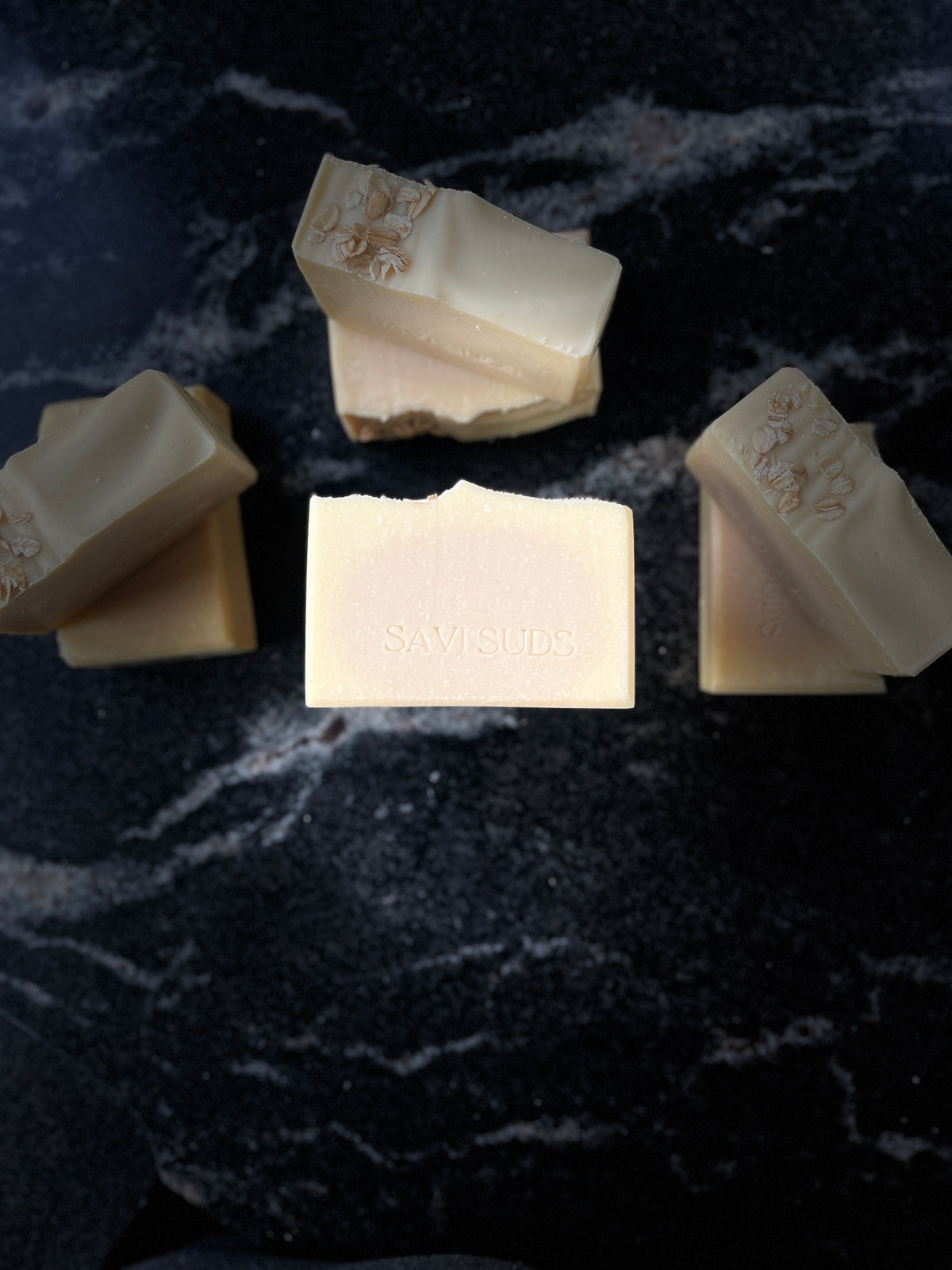 Seven bars of SAVI SUDS soap arranged on a dark, textured black marble surface, with one bar centered and facing forward to show the stamped brand name.