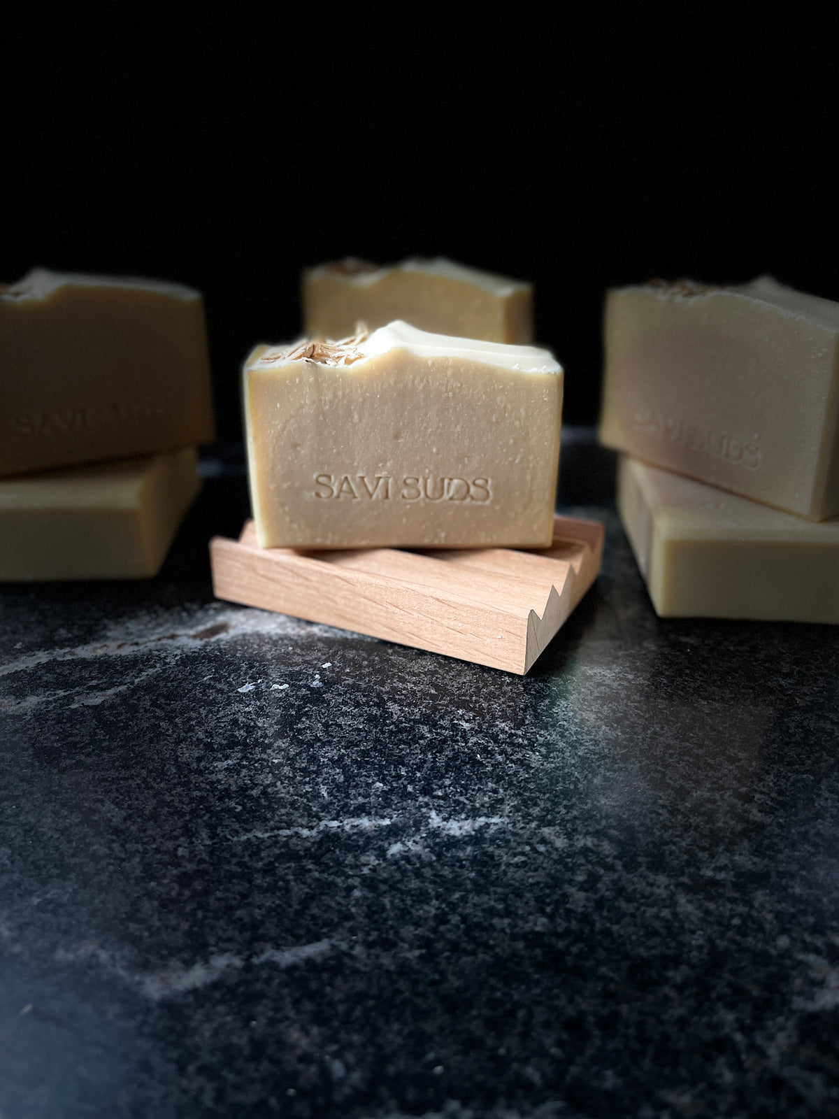 Several natural soap bars in a warm cream-beige color are arranged on a dark stone surface, with one central bar resting on a small wooden block. The front bar is stamped with the brand name 'SAVI SUDS'.