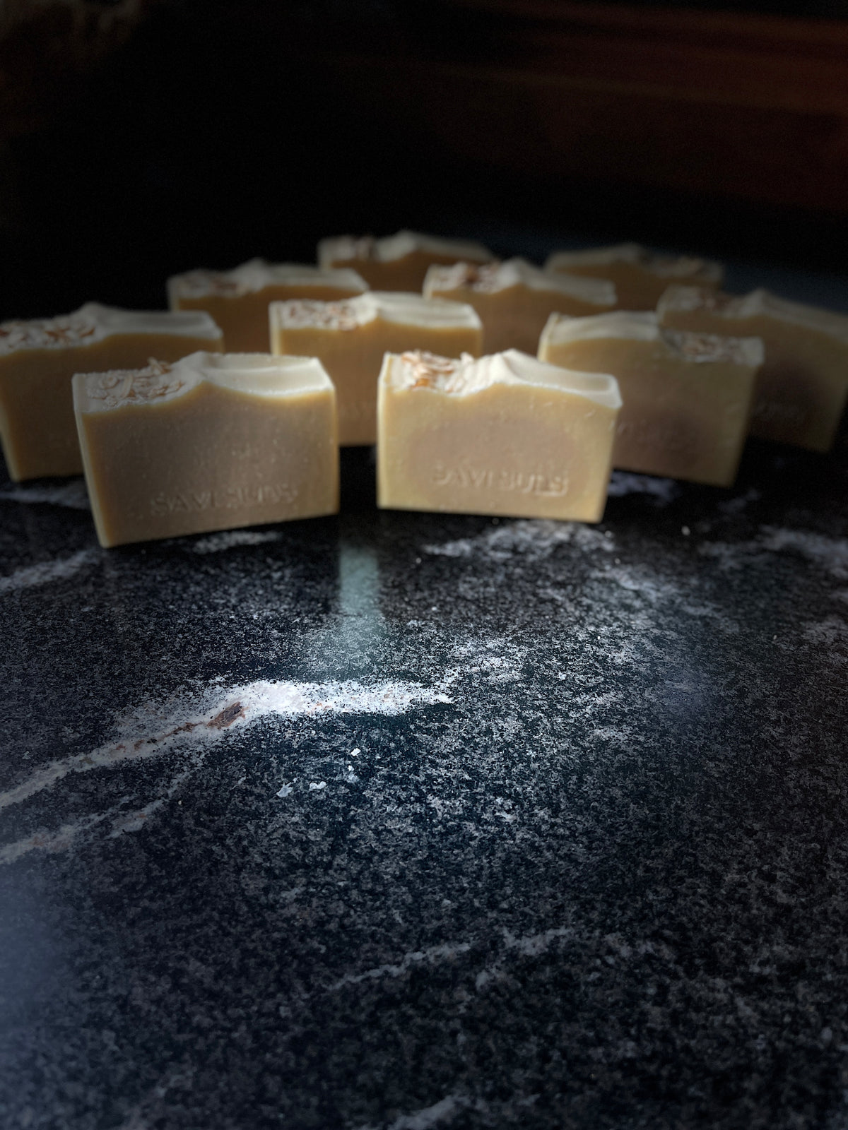 Several rectangular bars of pale beige soap with SAVI SUDS stamped on the front, arranged on a dark marble-like surface with a shallow depth of field.