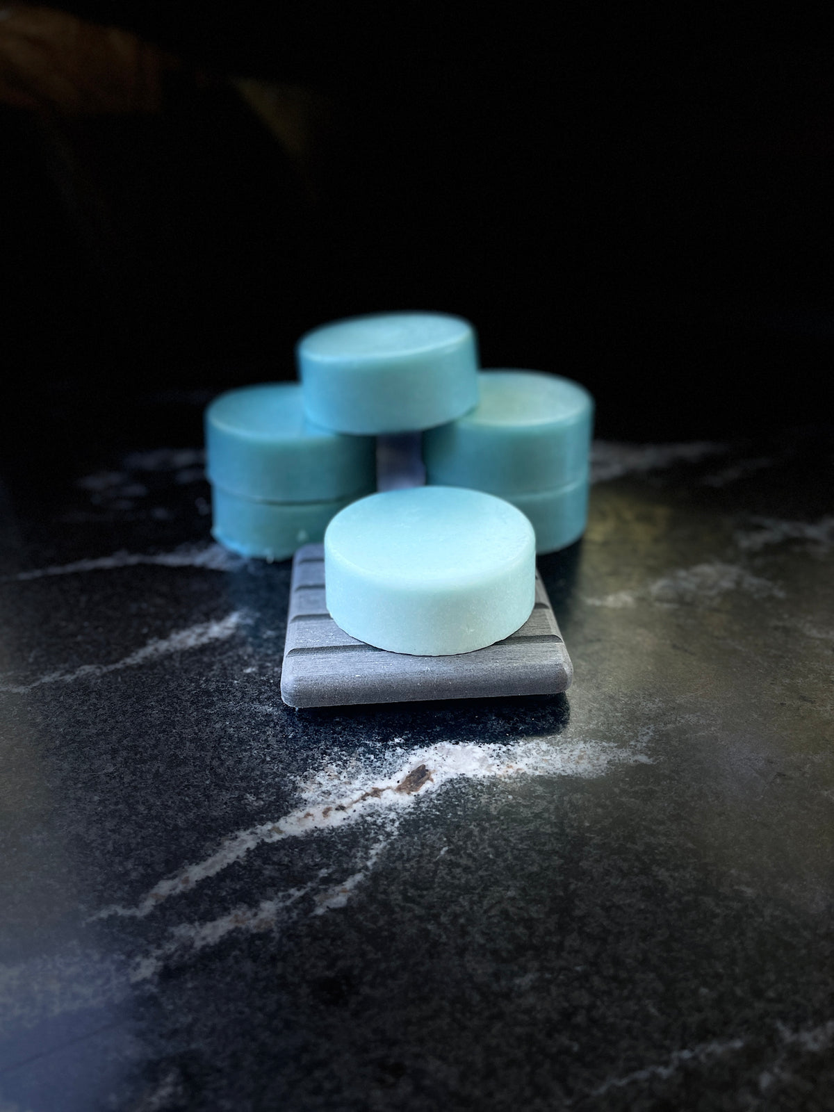Six ocean orchid round blue conditioner bars arranged on a dark marbled surface, with one bar resting on a gray soap dish in the foreground