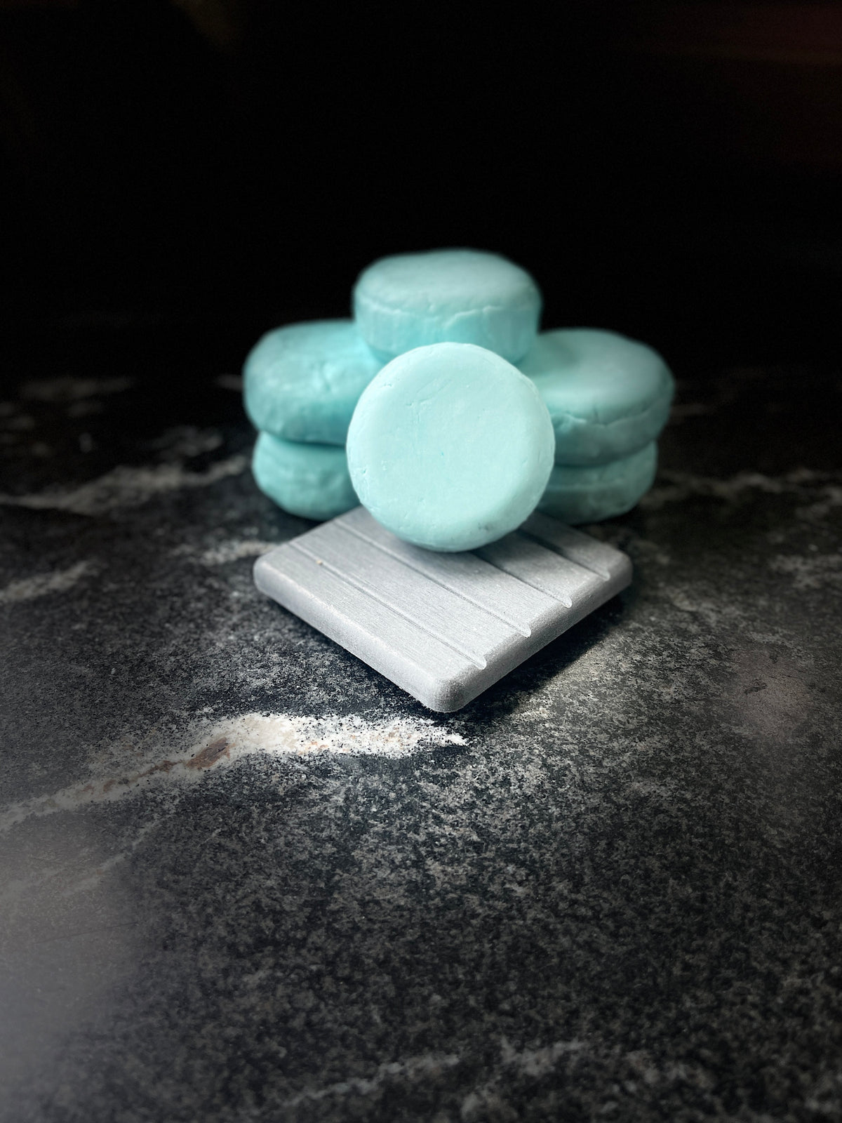 A stack of round, light blue Ocean Orchid shampoo bars arranged on top of a square grey stone, set against a dark, textured background.