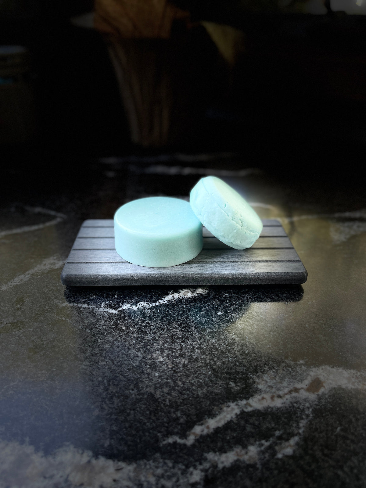 Two round pale blue soap bars resting on a small dark gray wooden slatted tray, set on a dark reflective countertop with a blurred background.