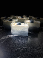 A bar of Savi Suds Onyx soap in the foreground, with several identical bars blurred in the background. The soap has a cream-colored top with lined throughout and a dark gray to black gradient base, topped with a small dark onyx soap.