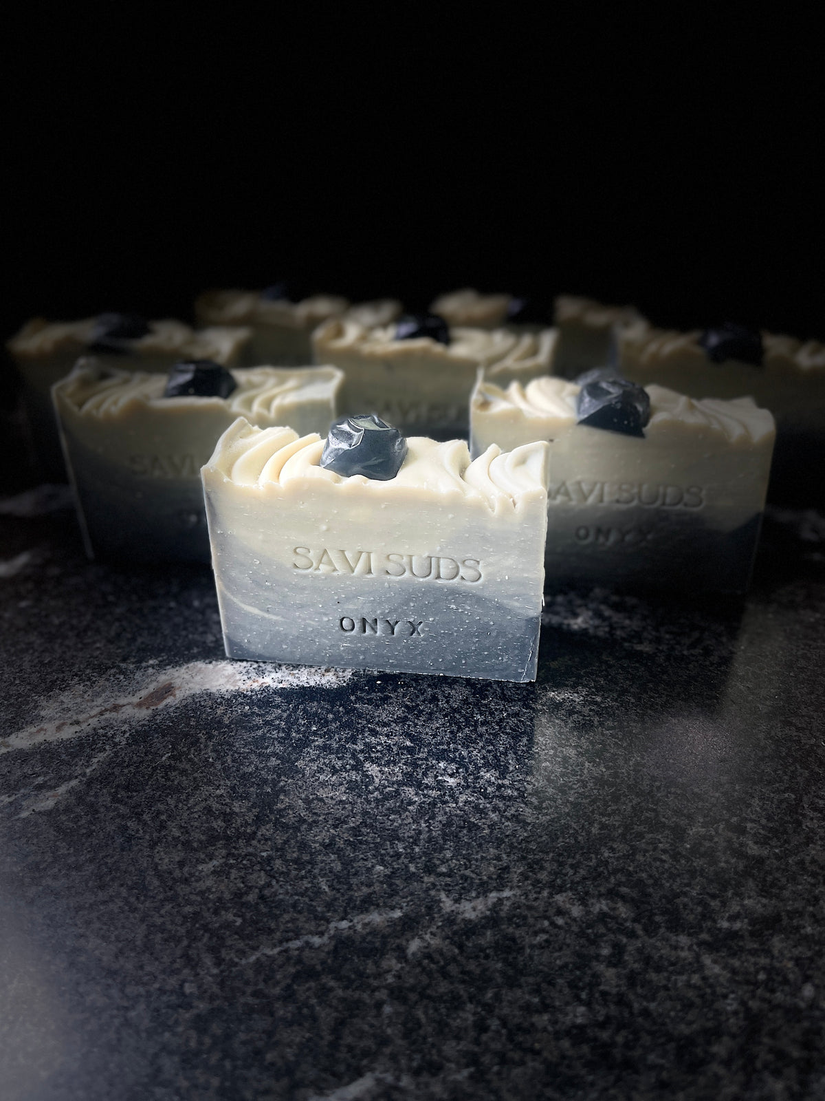 A bar of Savi Suds Onyx soap in the foreground, with several identical bars blurred in the background. The soap has a cream-colored top with lined throughout and a dark gray to black gradient base, topped with a small dark onyx soap.