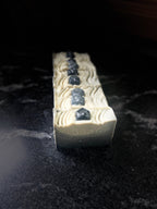 A line of rectangular soap with a textured cream-colored surface, decorated with a vertical row of dark soap stones, standing upright on a dark, reflective surface.