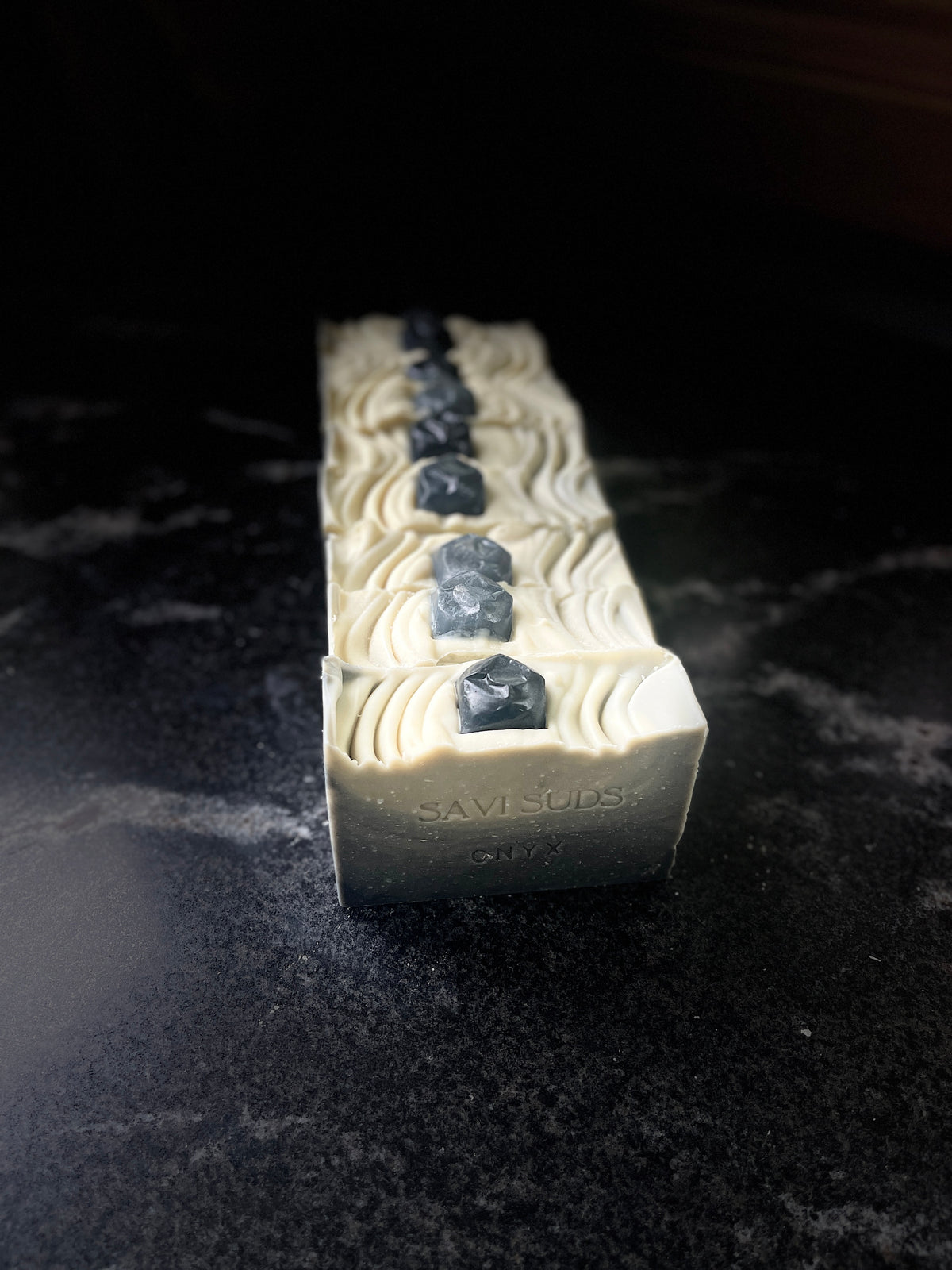 A line of rectangular soap with a textured cream-colored surface, decorated with a vertical row of dark soap stones, standing upright on a dark, reflective surface.