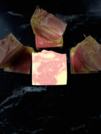 Several artisanal soap bars with pink and pale yellow marbled layers and dried botanical inclusions, arranged on a dark marble surface. The central bar is embossed with the brand name 'Savi Suds' and 'Orange Bergamot'.