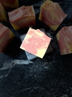 Several square bars of marbled pink and pale yellow soap are arranged on a dark stone surface. One soap bar in the foreground is slightly tilted and shows the embossed brand name 'SAVI SUDS' along with 'ORANGE BERGAMOT'.