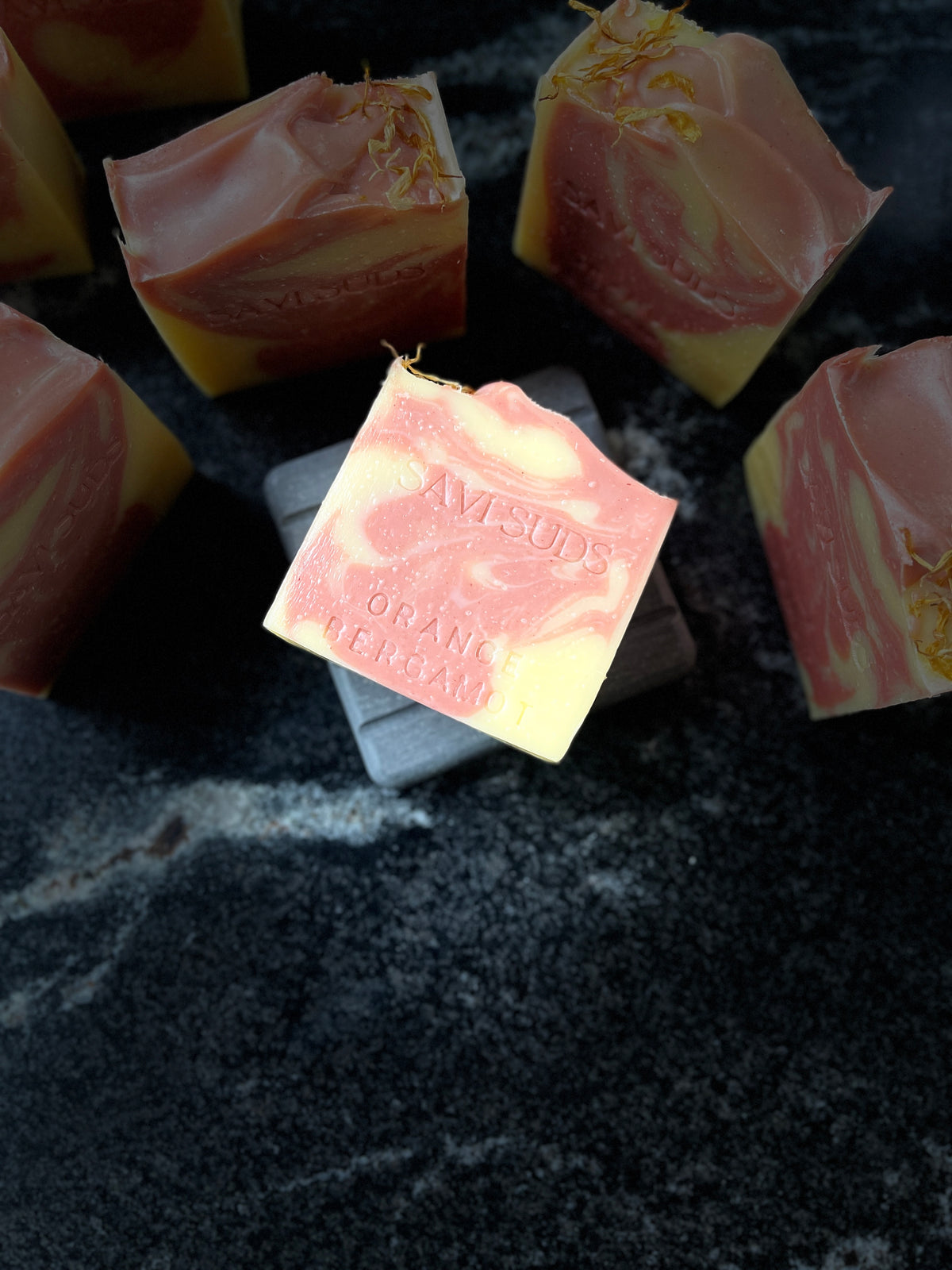Several square bars of marbled pink and pale yellow soap are arranged on a dark stone surface. One soap bar in the foreground is slightly tilted and shows the embossed brand name 'SAVI SUDS' along with 'ORANGE BERGAMOT'.