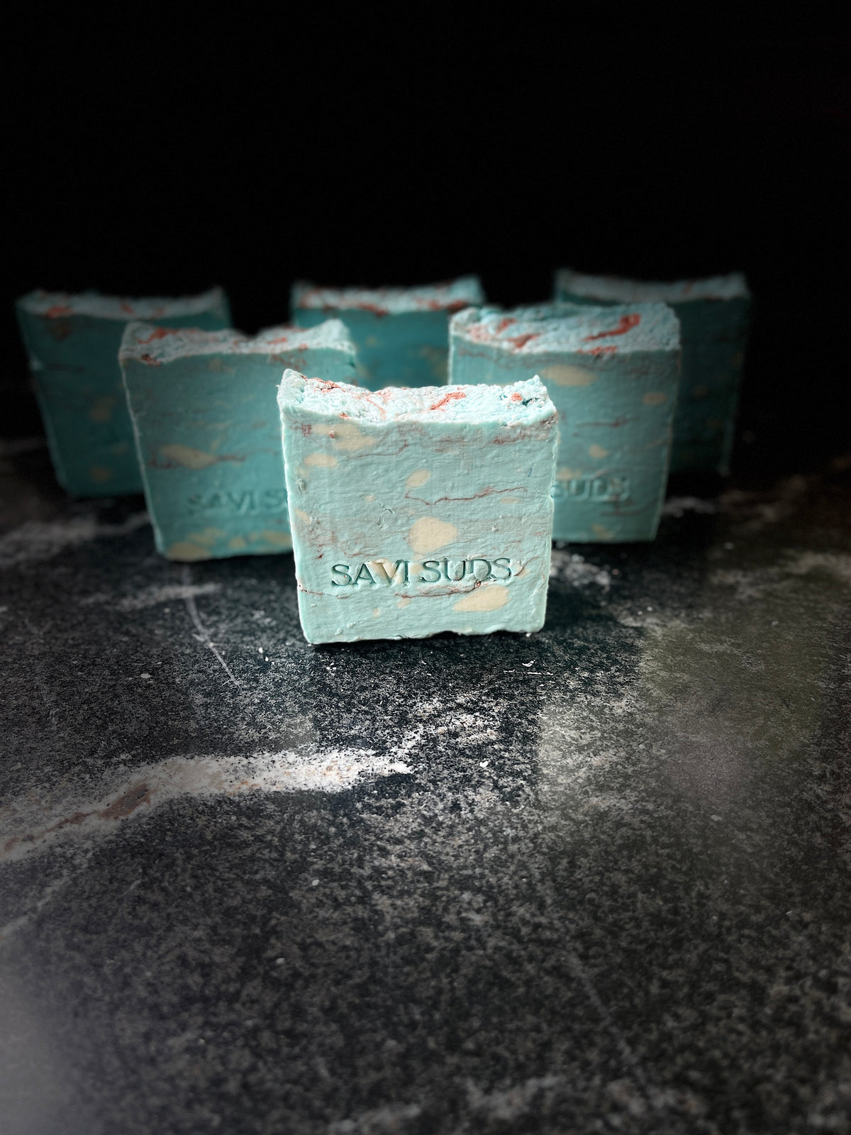 Several square bars of Savis Suds soap in a pale blue color with small white blobs and cider lines and spots, arranged on a dark textured surface.