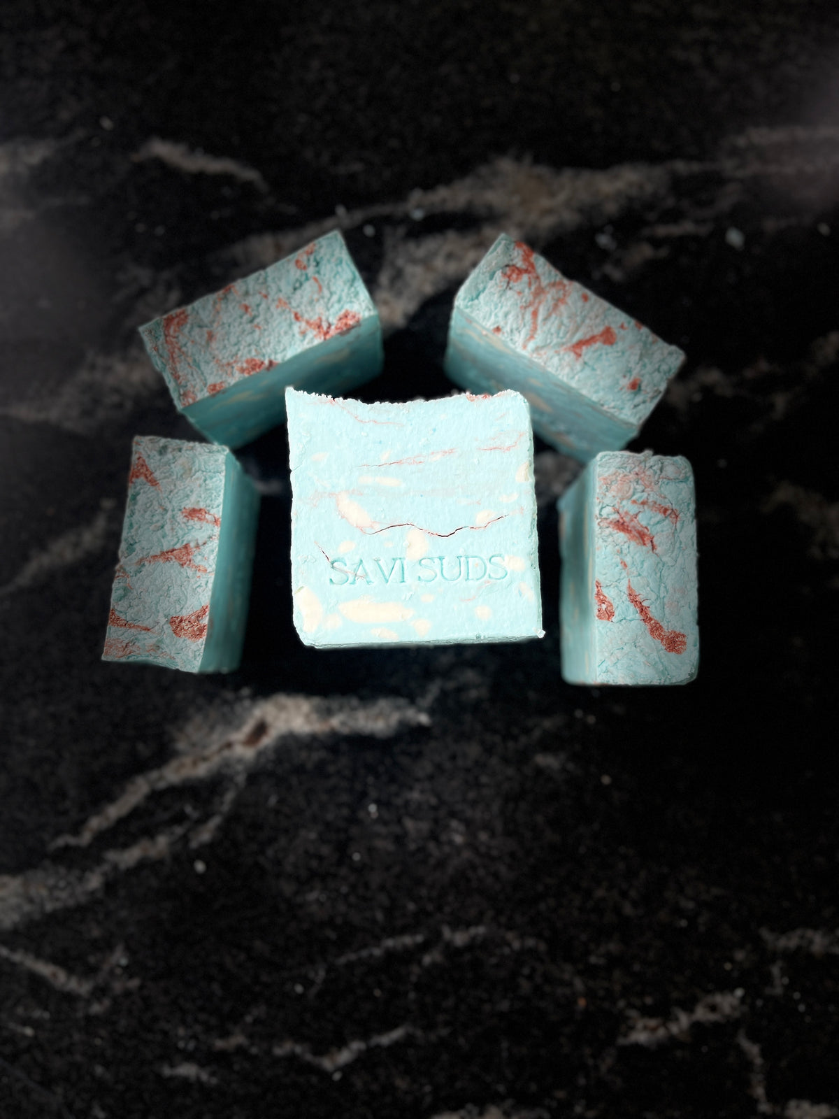 Five Square bars of light blue soap with orange-red marbled streaks, arranged on a dark marble surface. The center bar is embossed with the brand name 'SAVI SUDS'.