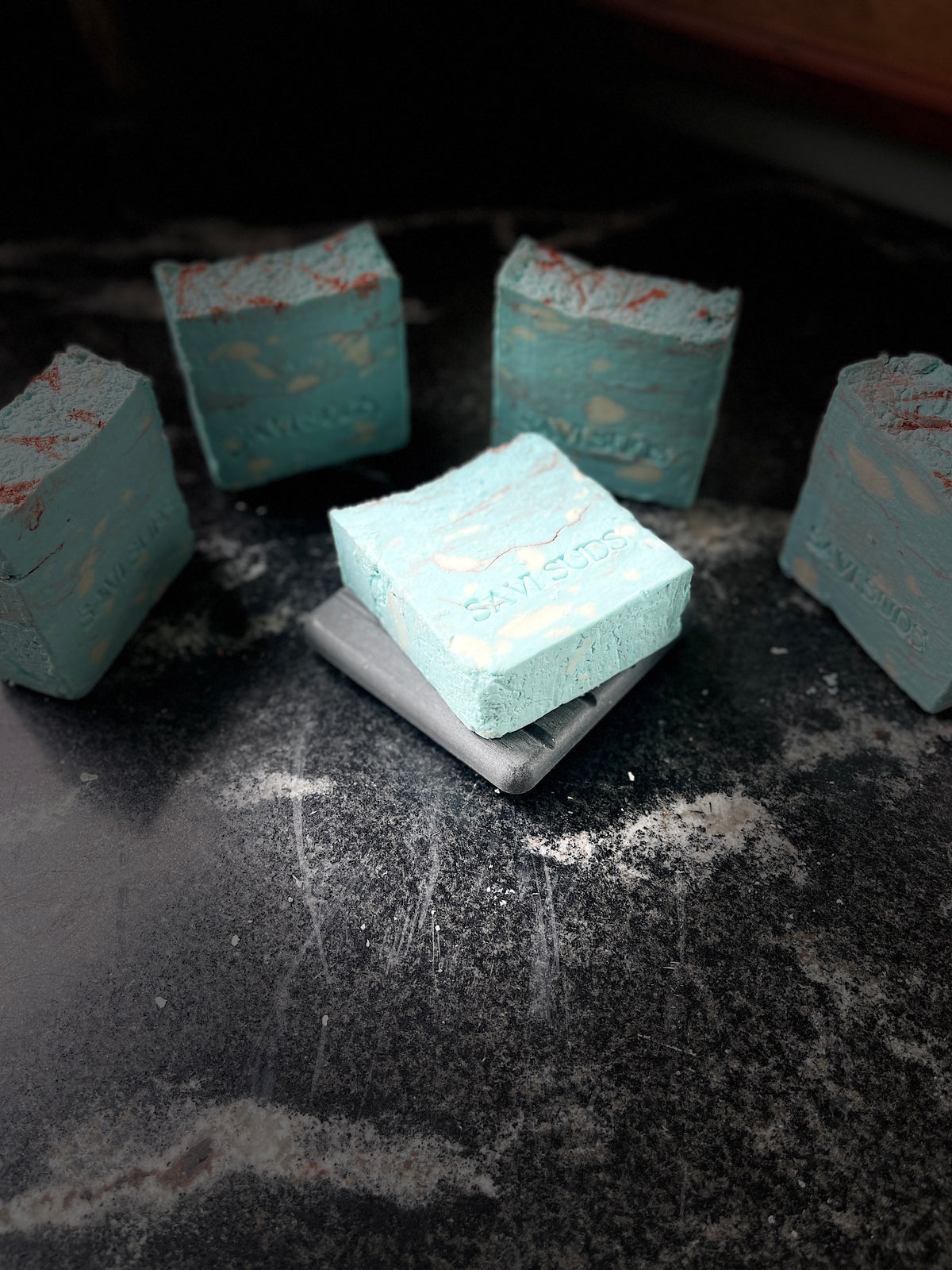 Five square bars of handmade soap in a light aqua blue color with white marbling and reddish-brown speckles, arranged on a dark, textured surface. One bar is placed on top of a gray square block, and the soap bars have the brand name 'SAVI SUDS' embossed on them.