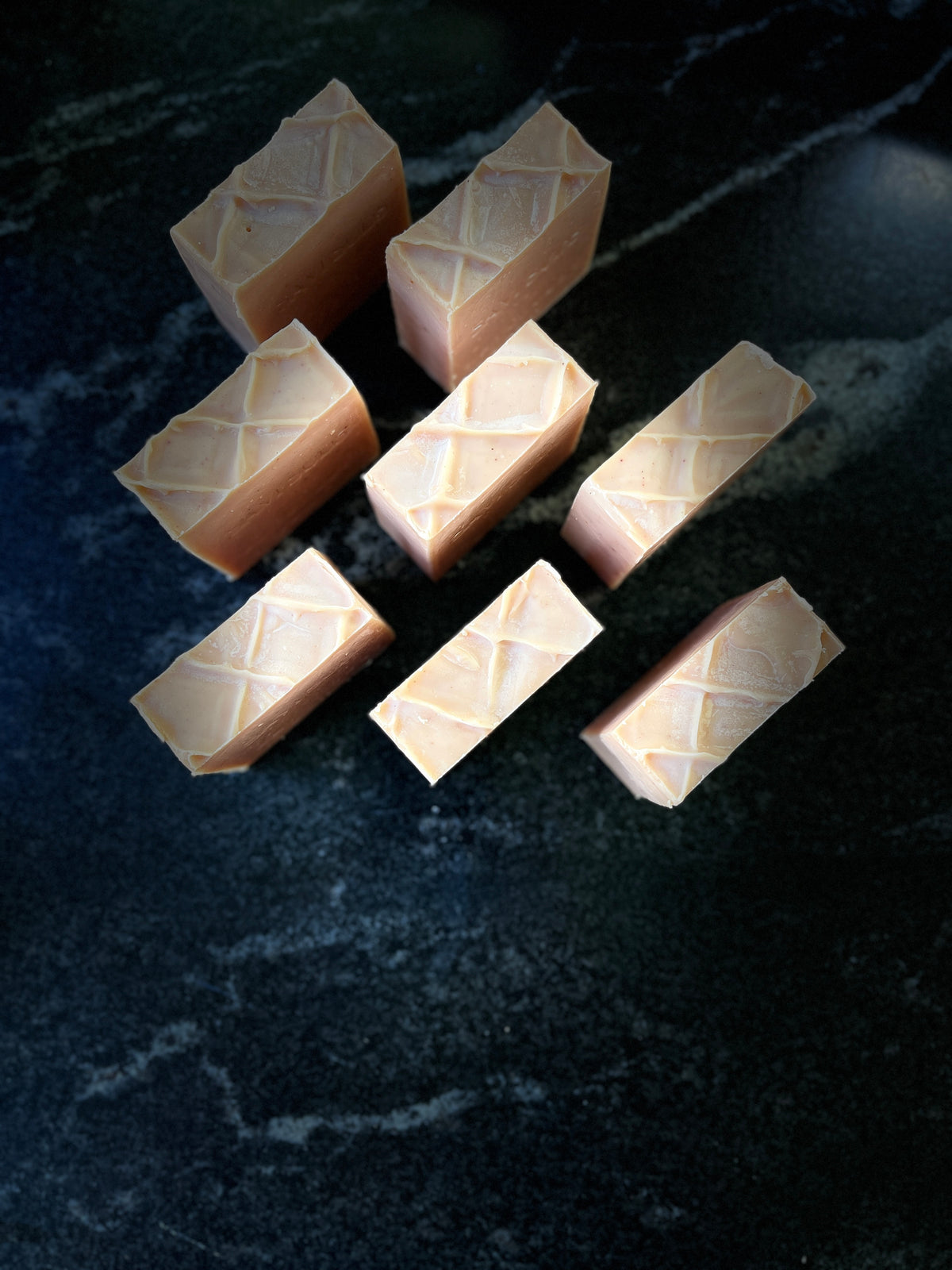 Eight square bars of pale peach-orange colored soap with a diamond embossed pattern, arranged on a dark textured surface.