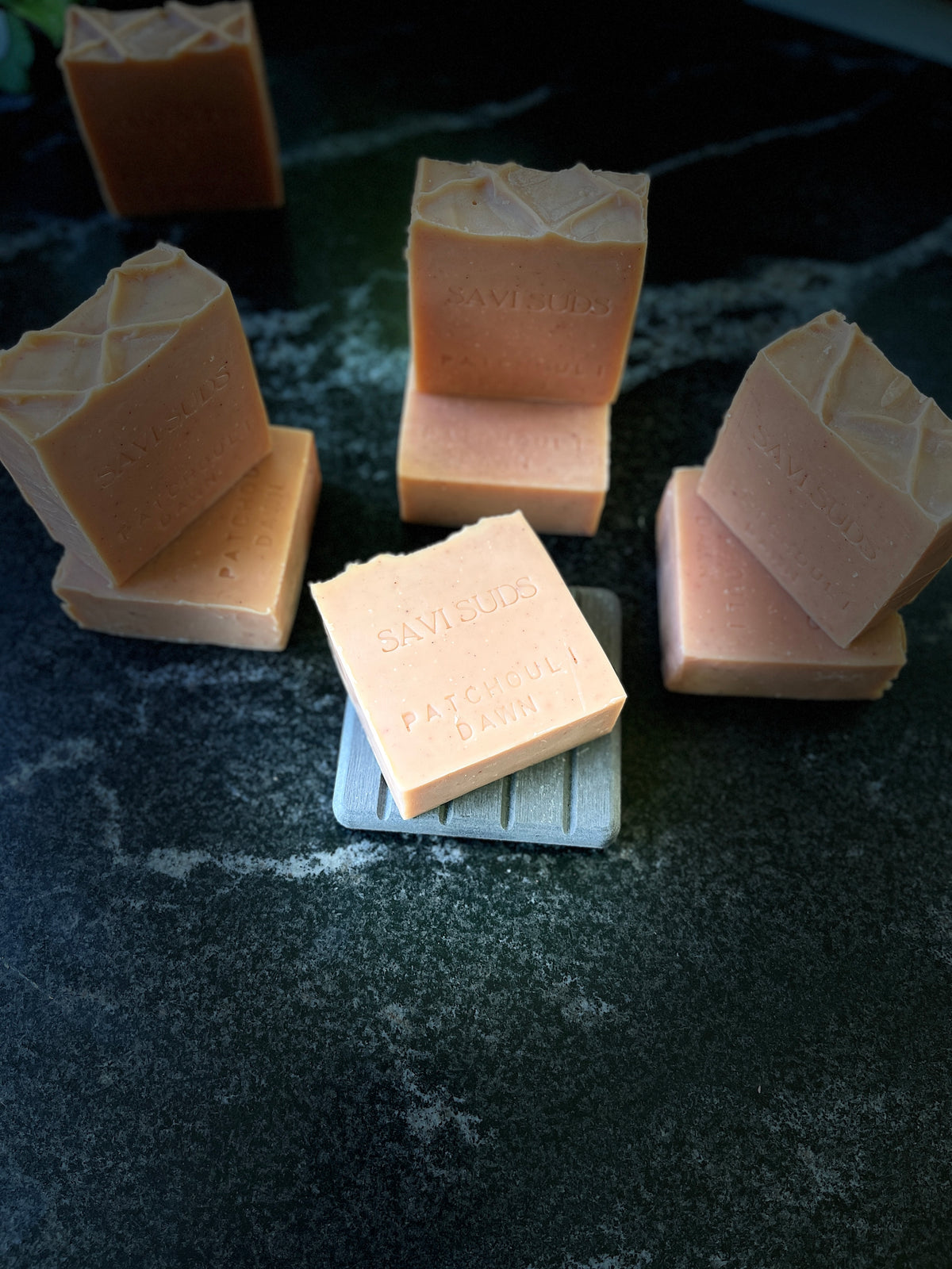 Several pale peach-orange colored handmade soap bars stacked on a dark textured surface. One soap bar in the foreground is stacked on a small white square and stamped with the brand name 'Savi Suds' and Patchouli Dawn