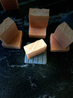 Several square bars of handmade soap in a warm peach-orange color, arranged on a dark textured surface. One soap bar in the center sits on a small grey slated soap dish