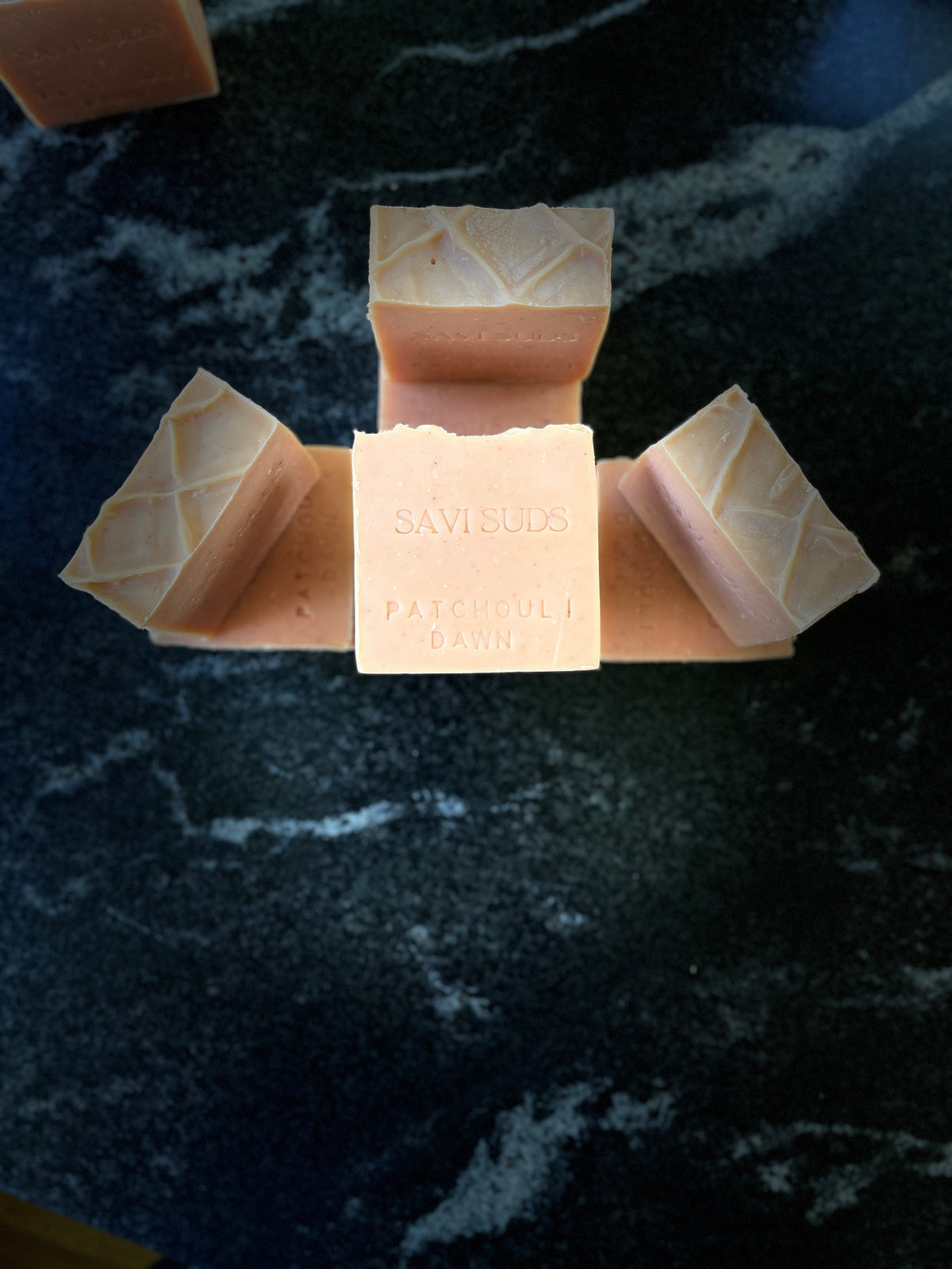 Several square bars of Savi Suds Patchouli Dawn handmade soap arranged on a dark marble surface, with one bar centered and the others fanned out around it.