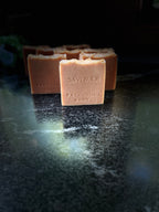 A handmade square bar of soap labeled 'SAVI SUDS PATCHCOULI DAWN' in a warm peachy-orange color, standing on a dark reflective surface with other bars blurred in the background.
