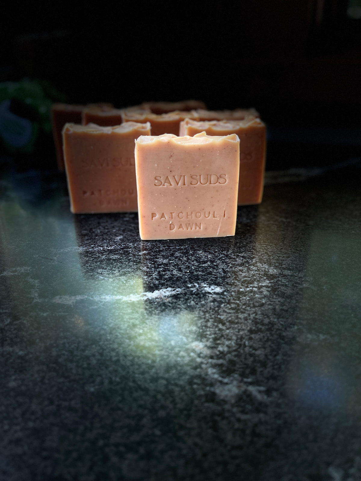 A handmade square bar of soap labeled 'SAVI SUDS PATCHCOULI DAWN' in a warm peachy-orange color, standing on a dark reflective surface with other bars blurred in the background.