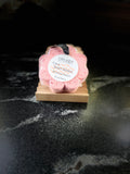 A pink flower-shaped bath soap sponge on a small wooden soap dish, with a round white label reading 'Savi Suds' and 'Soapy Sponge'. The dish sits on a dark marble-like surface against a dark background
