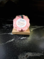 A pink flower-shaped bath soap sponge on a small wooden soap dish, with a round white label reading 'Savi Suds' and 'Soapy Sponge'. The dish sits on a dark marble-like surface against a dark background