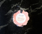 A pink flower-shaped bath sponge by Savi Suds, with a black ribbon and featuring a white circular label, set against a dark marbled background.