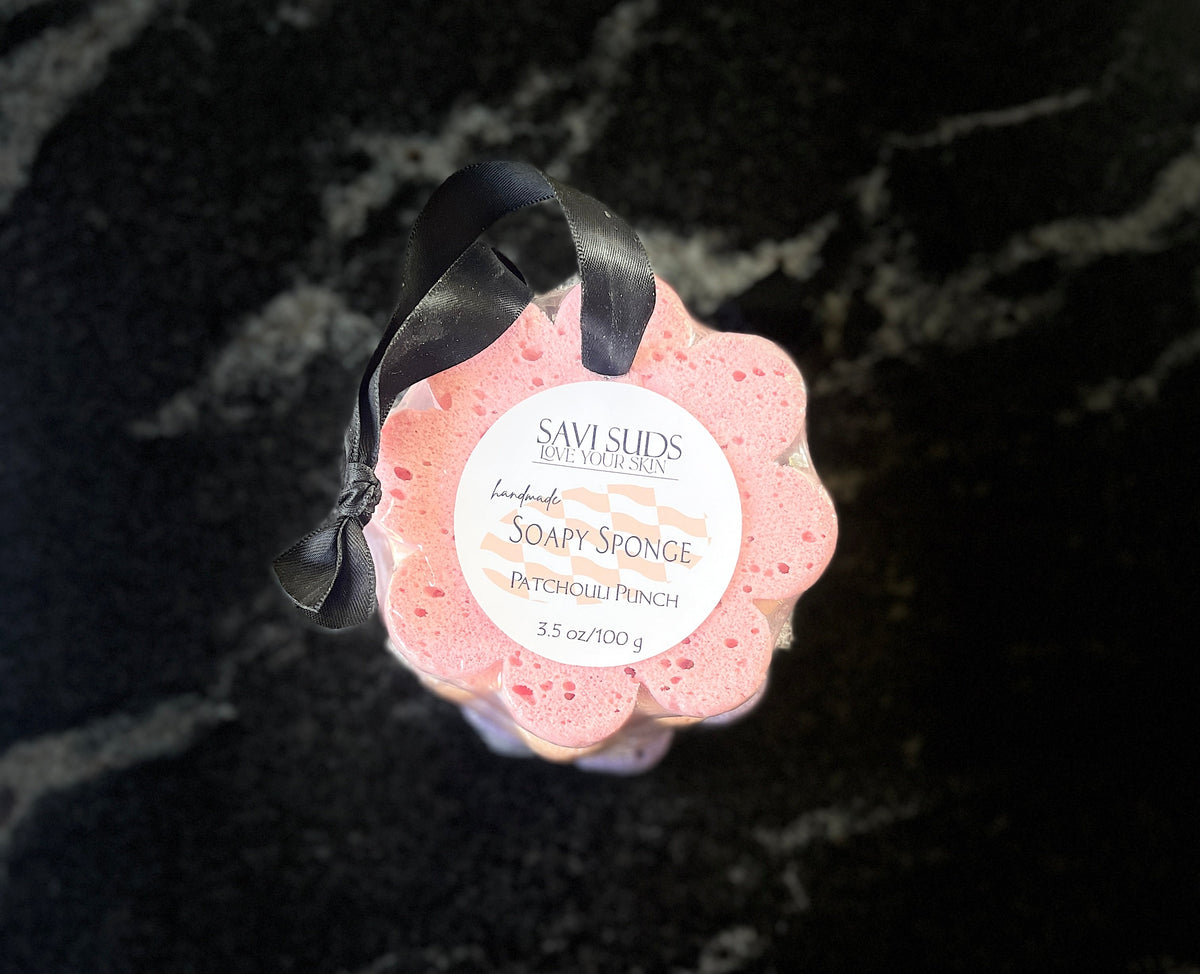 A pink flower-shaped bath sponge by Savi Suds, with a black ribbon and featuring a white circular label, set against a dark marbled background.