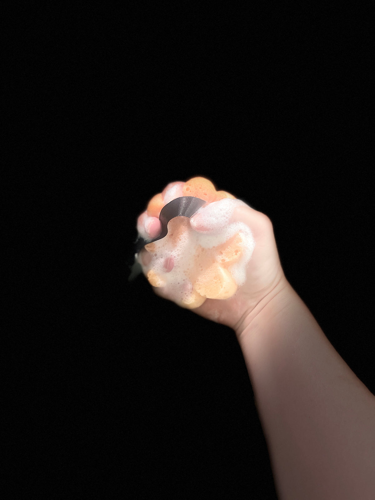 A hand squeezing a small, round, soap sponge against a solid black background.