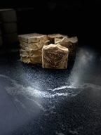 Handcrafted square bars of soap with marbled brown and cream swirls, stacked on a dark, textured surface with a soft spotlight effect.